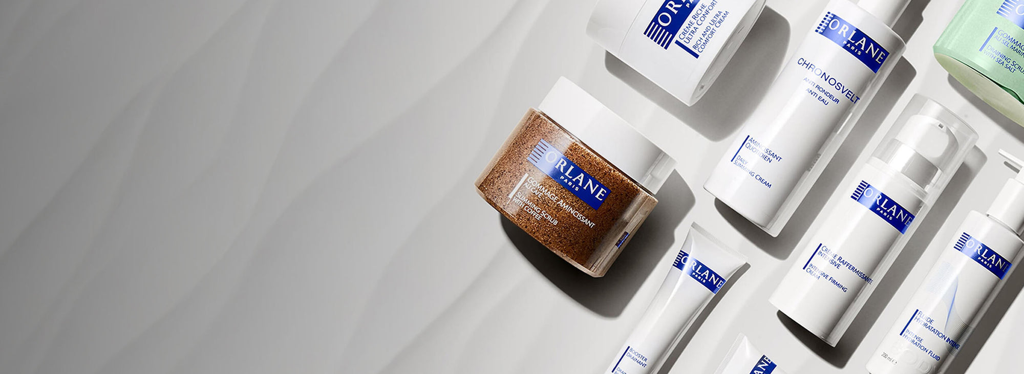 Flat lay of Orlane Paris body and slimming skincare products including slimming scrub, firming cream, and hydration fluid on a soft gray marble surface